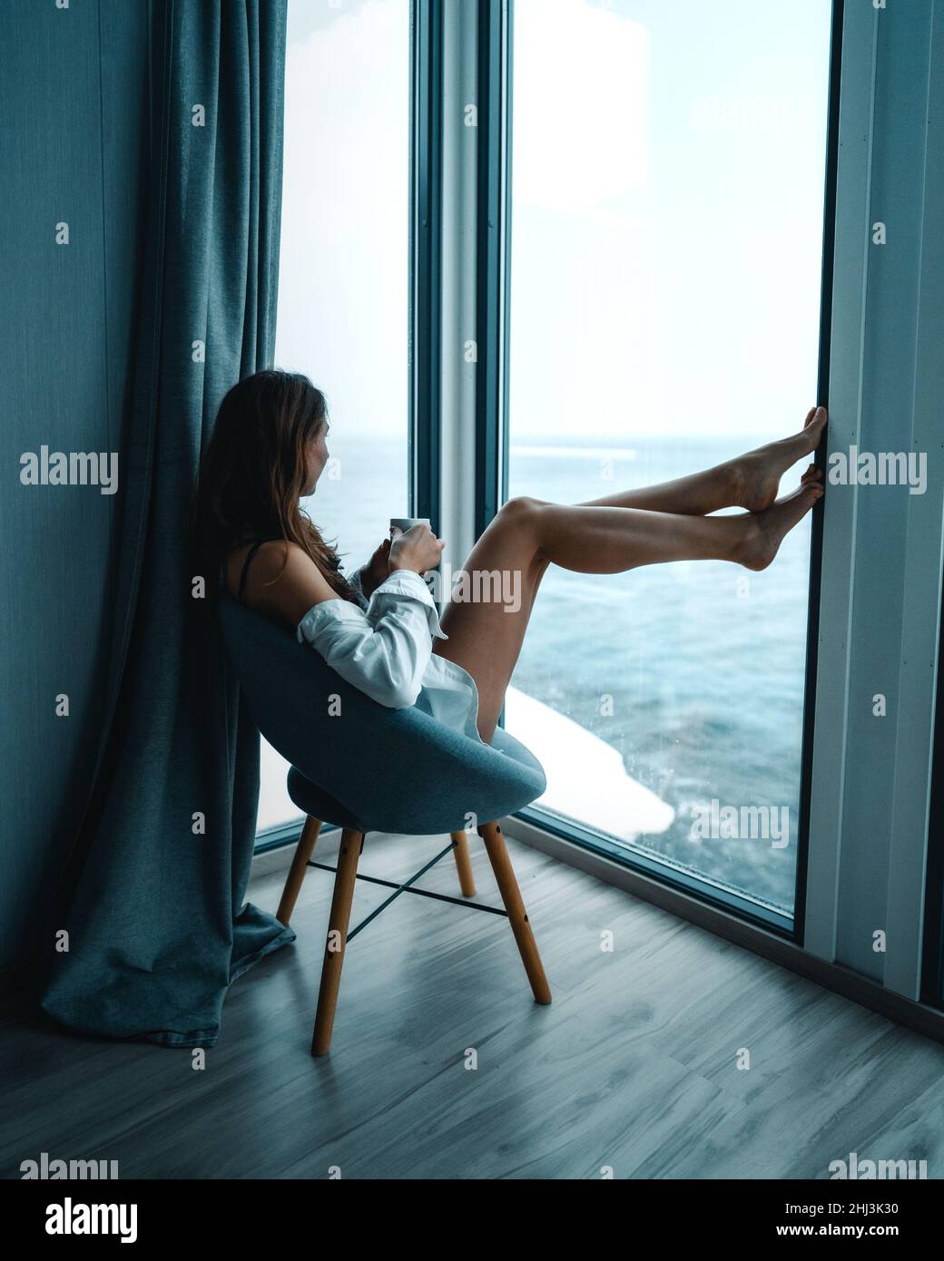 A young traveler women enjoying view with a morning coffee from a cabin by the ocean. Tiny house luxury travel in Norway in winter. Stock Photo