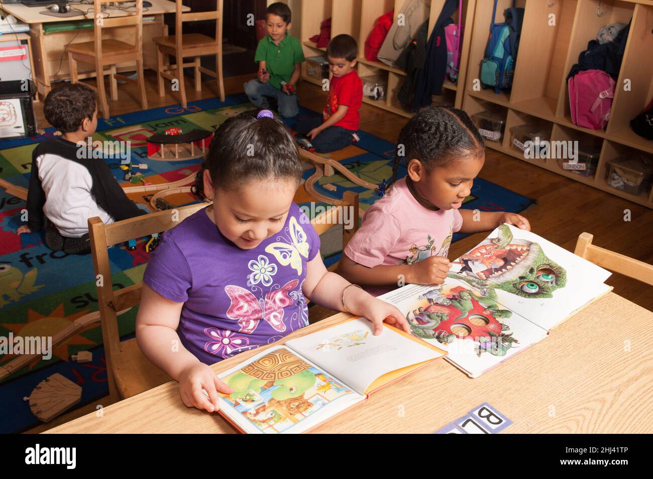 Education Preschool 4 year olds two girls sitting at table and looking quietly at books while group of boys play with train set and tracks in backgrou Stock Photo