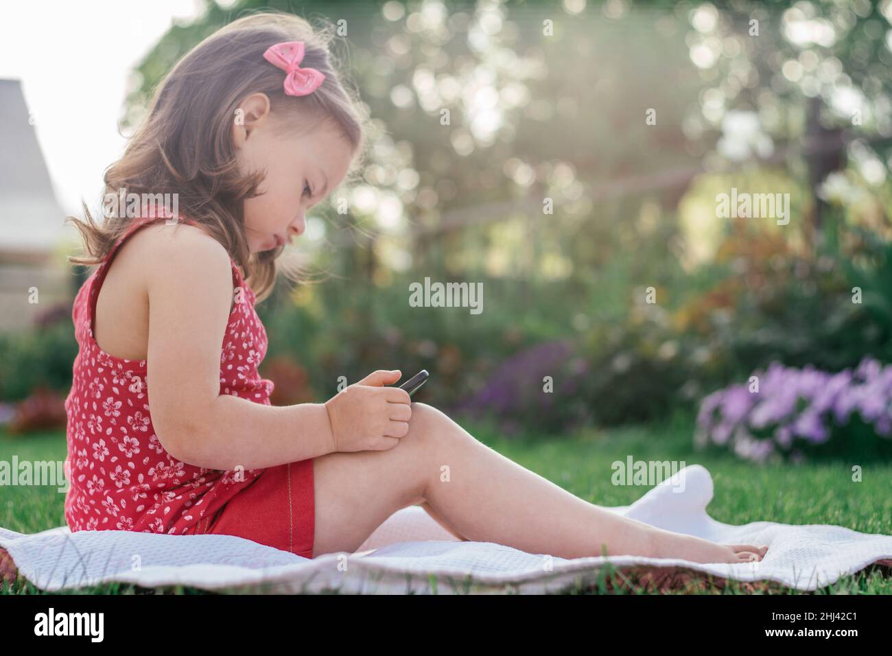 Little 34 girl in red clothes sits on blanket on green grass and looks