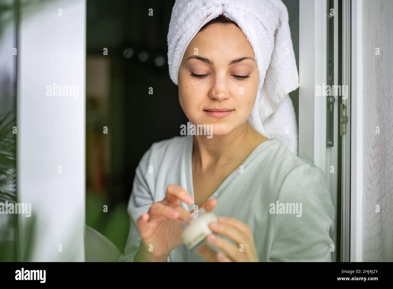 Happy domestic woman applying anti aging cream from jar on face looking at mirror Stock Photo