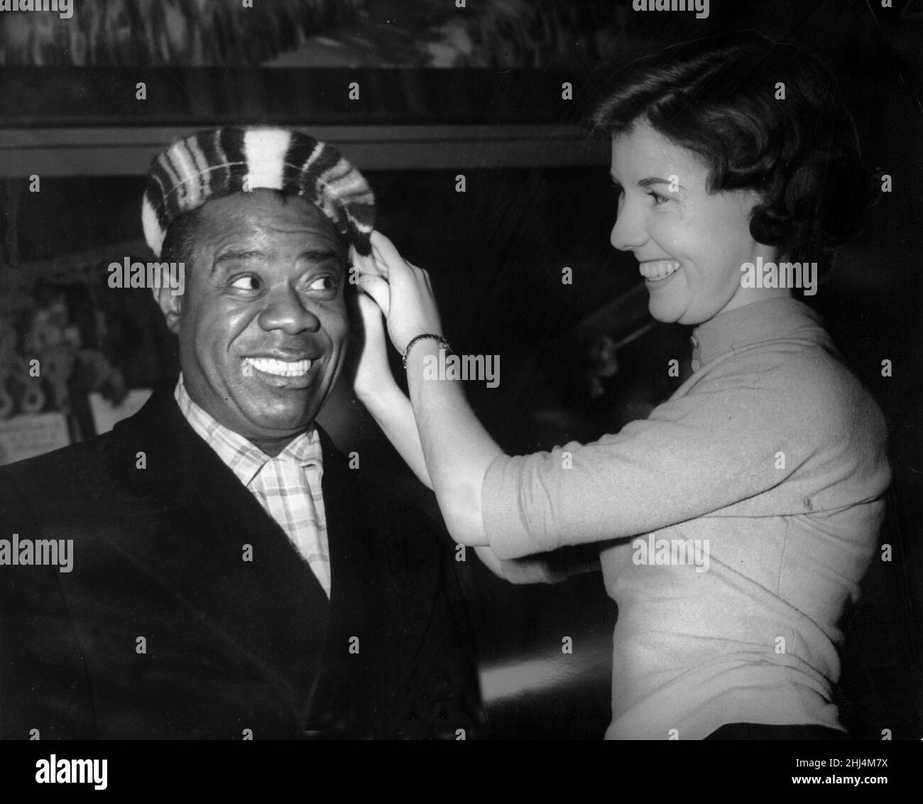 American jazz musician Louis Armstrong stops off at Prestwick Airport, Glasgow on his way back to the United States. Here he is having his beret fitted out by 24 year old May Halliday. 22nd December 1956. Stock Photo