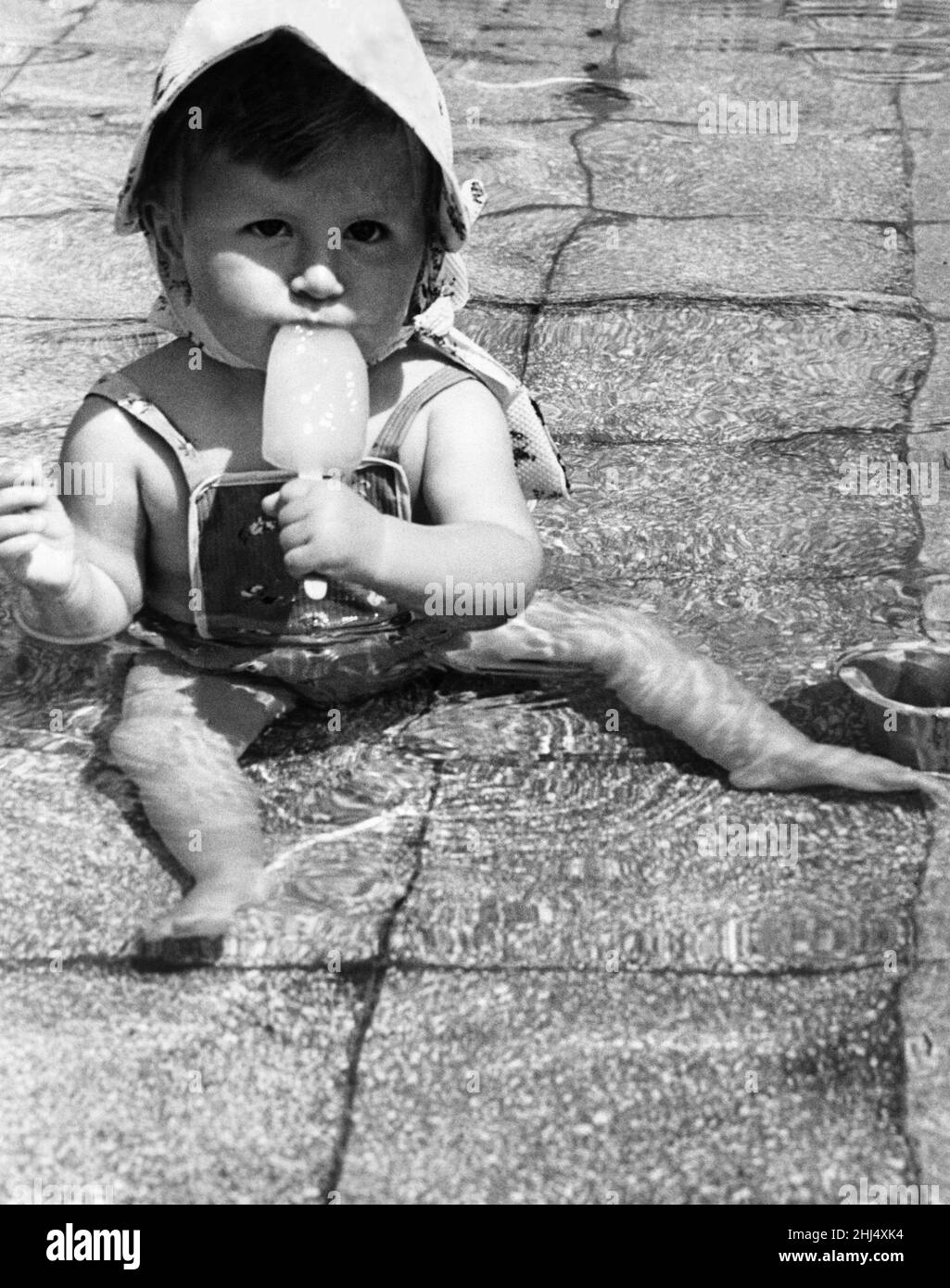 14 month old Lynda Hall from Burgess Hill, Sussex, decided the only way to keep cool yesterday (Tues) was to don a swimsuit and bonnet and sit down in the middle of the children's paddling pool at Black Rock in Brighton - and suck an iced lolly 9th July 1958. Stock Photo