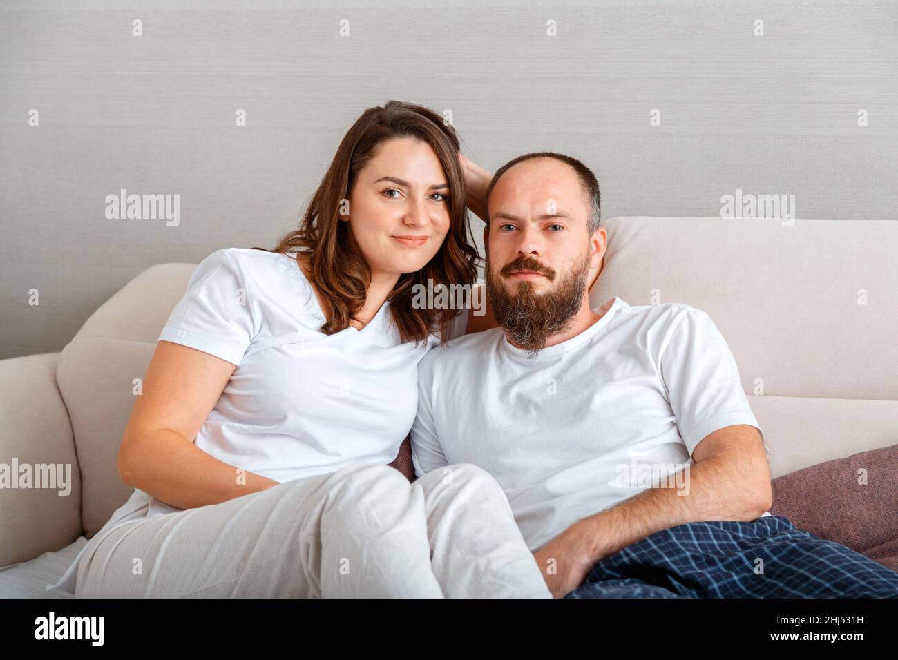 Young beautiful couple in pajamas hug while sitting on couch at cozy home interior. Married man and woman romantic relationship. Husband and wife Stock Photo