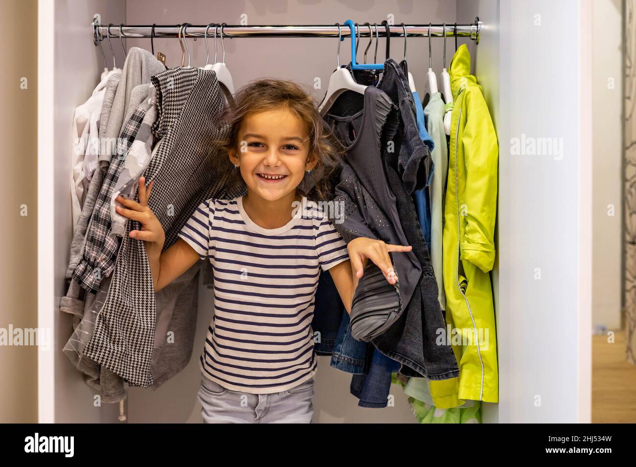 Portrait cute little girl posing hiding in cupboard with hanged clothes