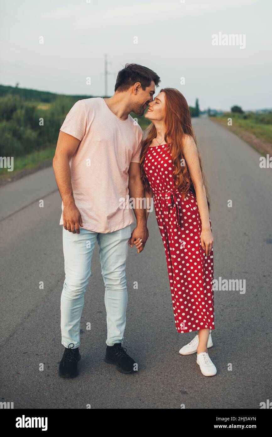 Full lenght of a man in a T-shirt and a redhead woman in a red jumpsuit with polka dots holding hands while traveling on an asphalt road. Summer Stock Photo