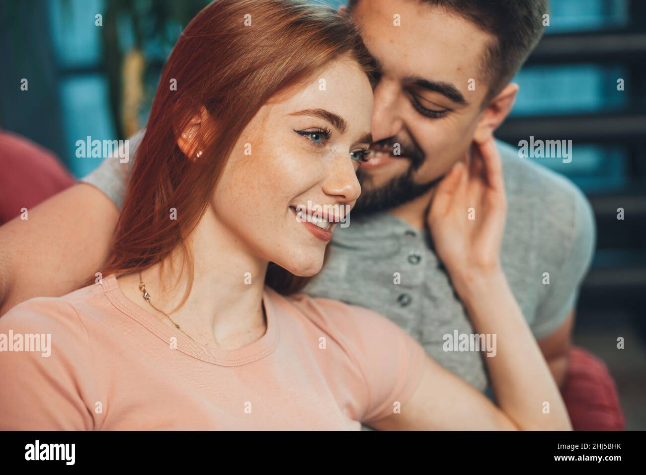 Close-up view of a Caucasian couple sitting on the couch on the weekend, freckled woman with red hair touching sensually her boyfriend's face. Stock Photo