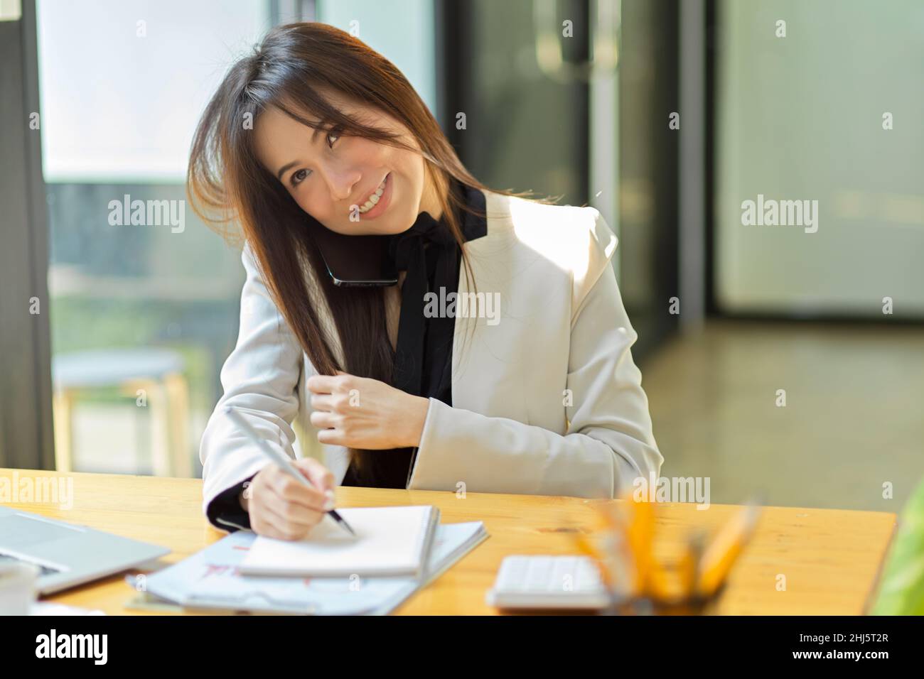 Attractive female assistant or secretary answering phone call and taking notes on her notepad at her office desk. Woman talking on the phone. Stock Photo