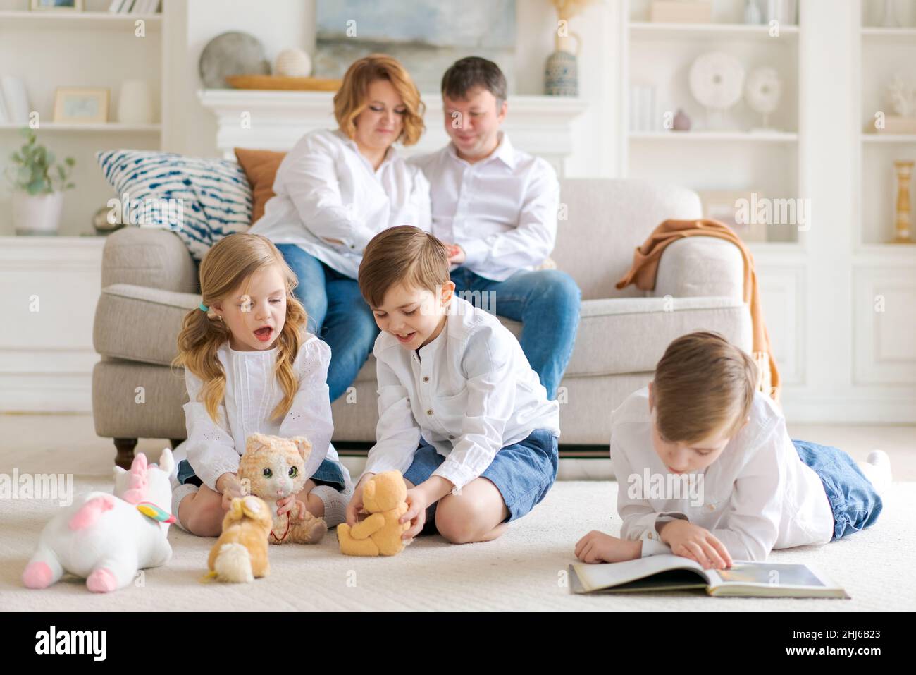 Happy young caucasian family resting in a new cozy design home on weekends. Delighted parents resting in their own home, children have fun sitting on the floor with toys and a book Stock Photo