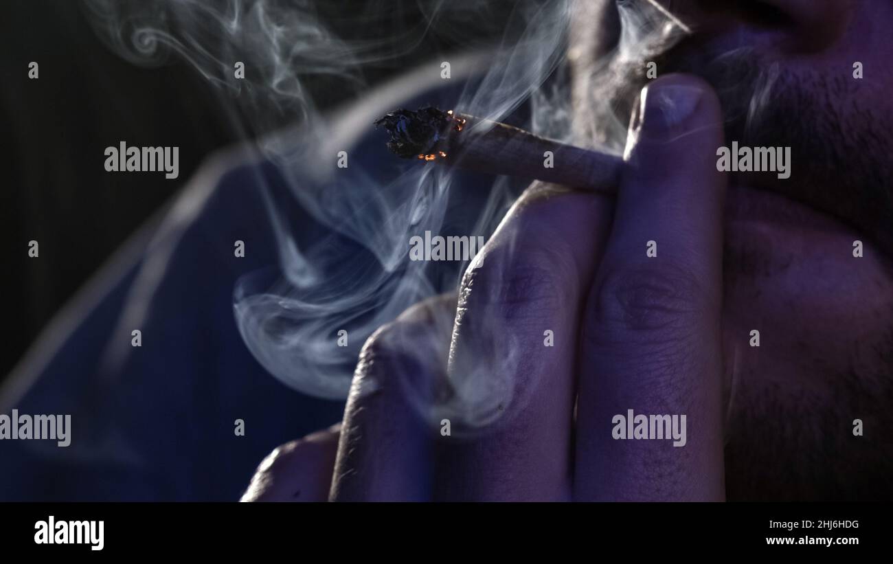 Male fingers with burning cannabis cigarette with smoke and cigarette glow under the blinking light, macro view. Stock Photo