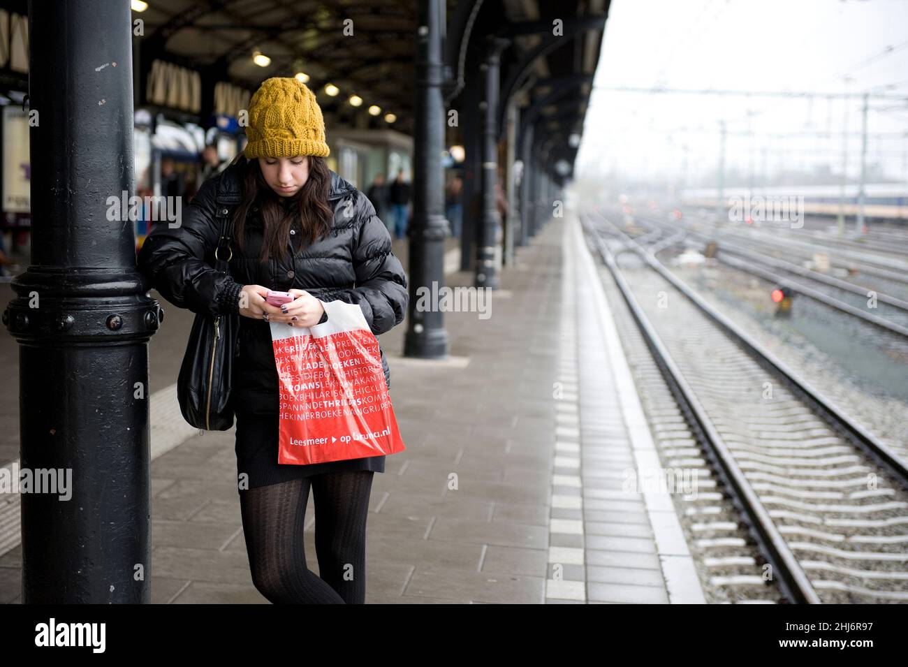 Den Bosch, 's-Hertogenbosch, Netherlands. Young adult woman leaning against a roof supporting pole on the local Railway Station while waiting for her connecting intercity train towards Breda. Stock Photo