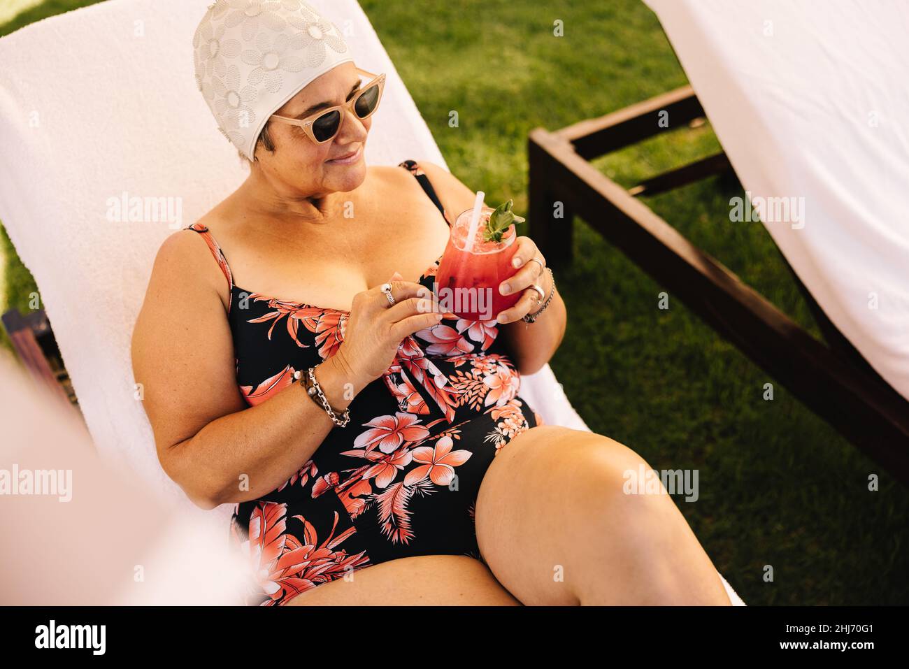 Mature woman having a tiki cocktail while relaxing on a lounger. Carefree elderly woman enjoying her summer vacation at a luxury spa resort. Senior wo Stock Photo