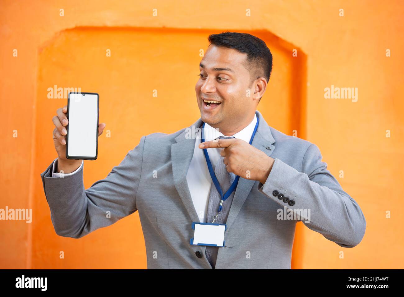 Excited young Indian man wearing suit and id card pointing at blank phone screen, Cheerful male manager hold empty smart phone display for advertiseme Stock Photo