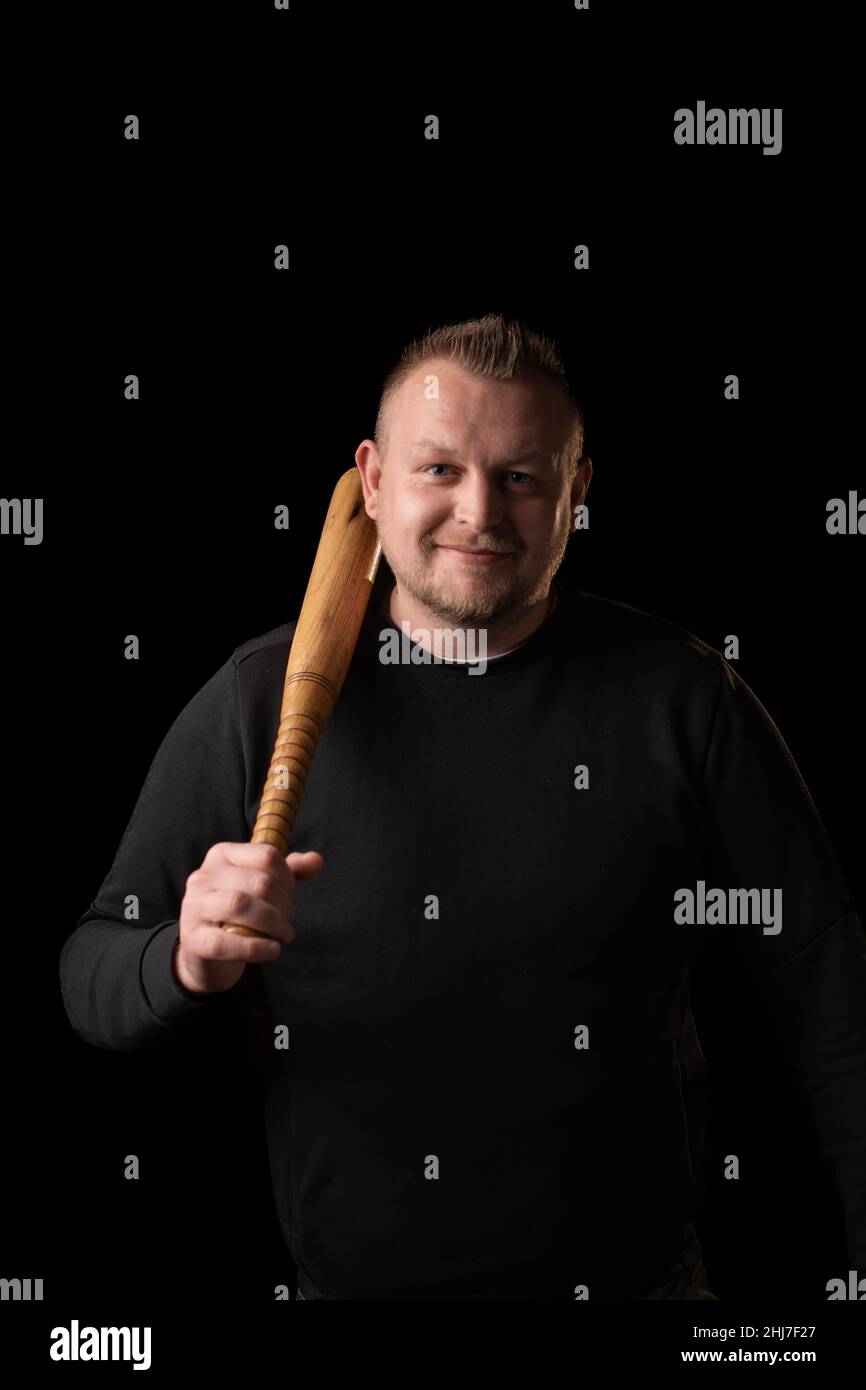 A young strong man holds a bat in his hand and smiles. Dark background. Man in black clothes. Bouncer, security guard, bodyguard concept. Stock Photo