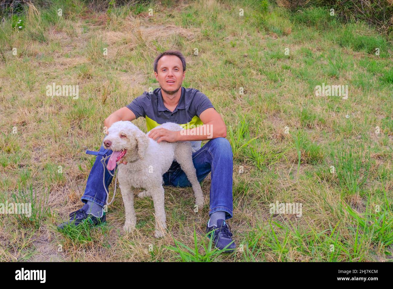 man with a white dog of a laggoto-Romagnolo breed sitting on grass in the park. Walking the dog in the park. dog in search of truffle mushroom Stock Photo