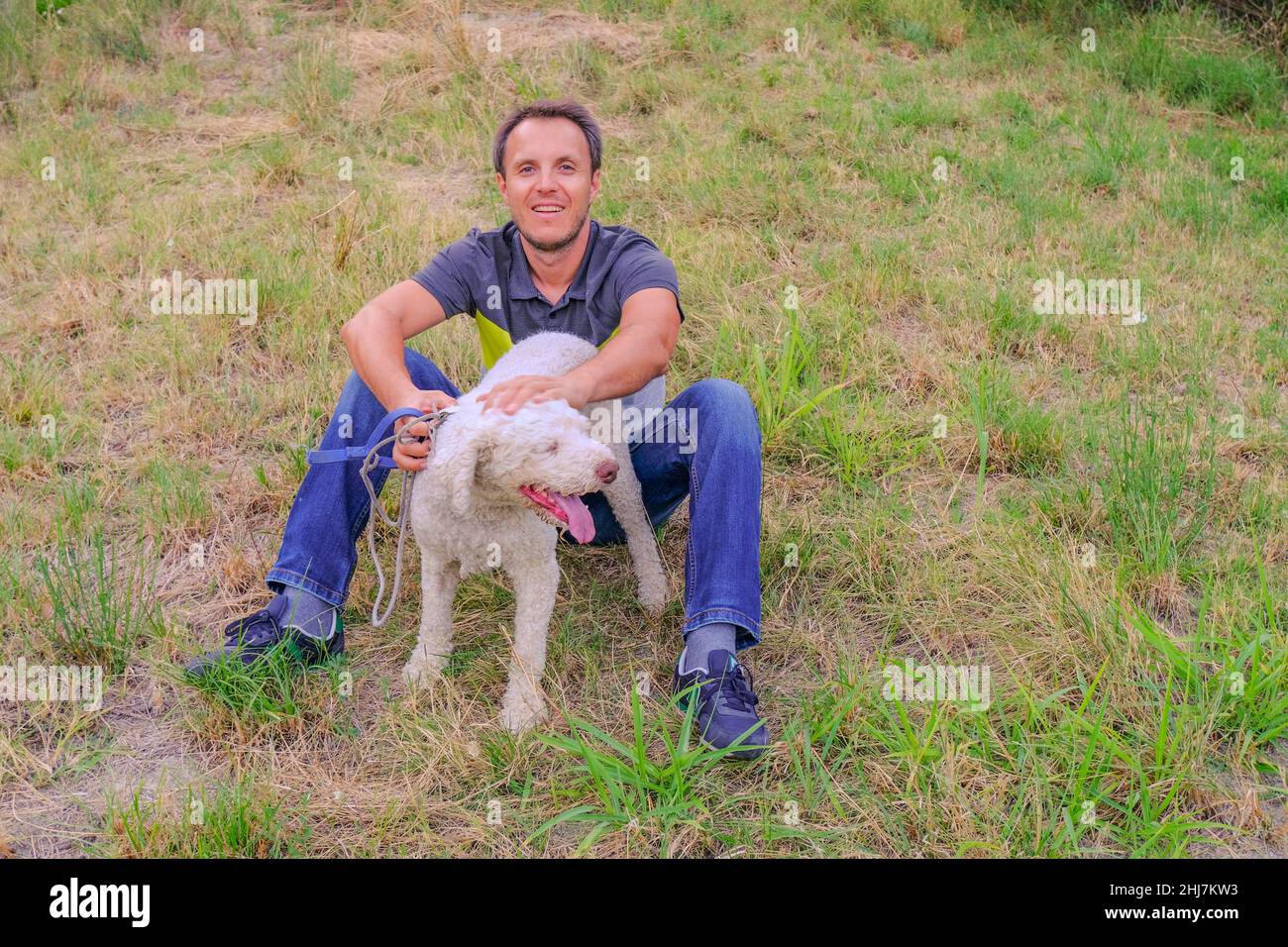man with a white dog of a laggoto-Romagnolo breed sitting on grass in the park. Walking the dog in the park. dog in search of truffle mushrooms Stock Photo