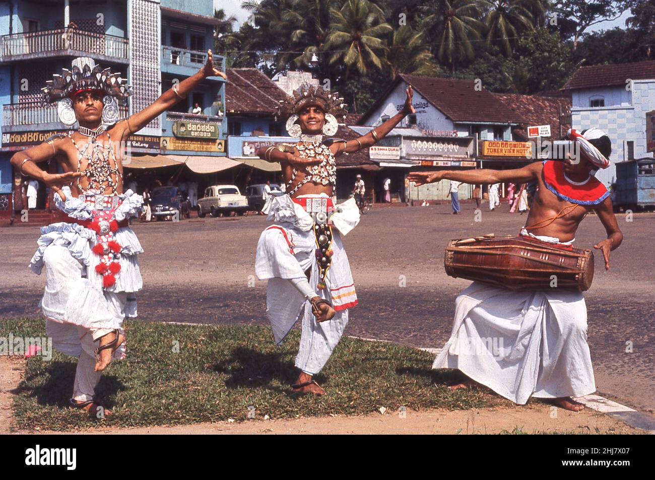 1960s, historical, Ceylon, outside in a village square, on a grass verge, two adult males in costumes, headwear  and decorated flowers, performing the traditional Kandyan dance, accompanied by a drummer playing the gata bera, the traditional srilankan drum. The Kandyan dance has a long history in the country and the ritual is performed with the belief that its blesses people with prosperity and long life. Since its orgins many centuries ago, the dance has become a symbol of Sri Lankan national identity. Stock Photo