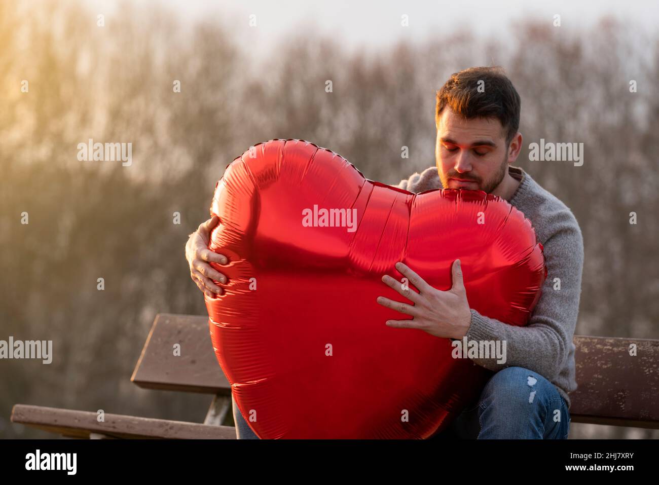 Sad young man hugging a heart-shaped balloon sitting on a bench in the park Stock Photo