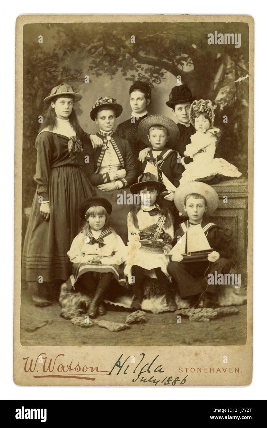 Lovely, clear, original Victorian era cabinet card studio portrait with painted backdrop, of large family group of women, teenage girls and children, lots of characters.  The young boys are dressed in fashionable sailor suits and are holding toy yachts and the young girl at the front, a basket of flowers (reinforcing gender stereotypes). Several nice hats and bonnets. The austere scary matriarch at the back is wearing an Order of Christ Cross (Portuguese Cross) brooch at her neck. From the studio of W. Watson, Stonehaven, Aberdeen, Scotland. On the front is written 'Hilda, July 1886'. Stock Photo