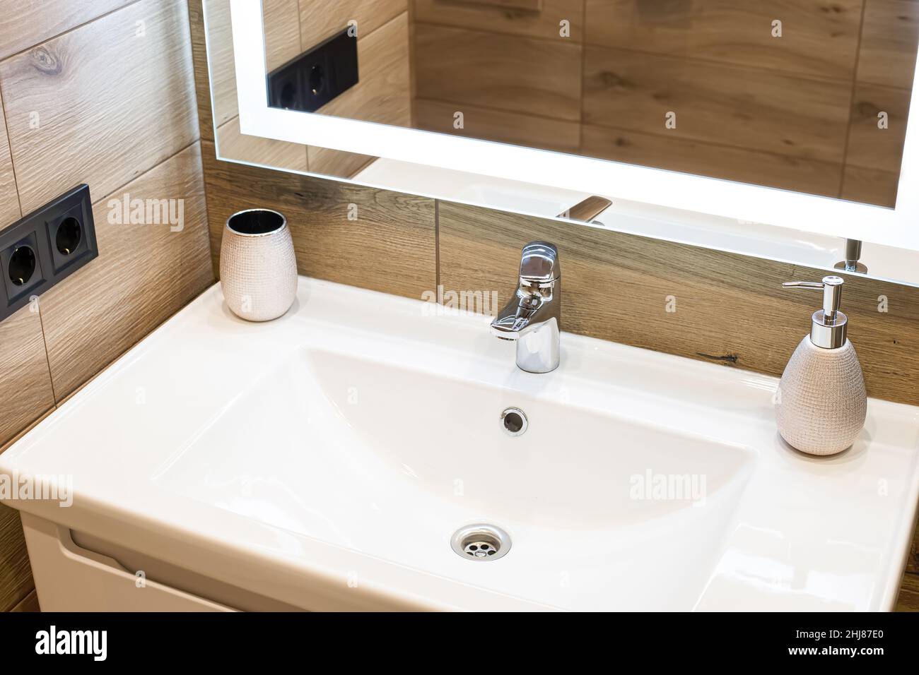 Modern washbasin with chrome faucet beside a stylish soap dispenser. Mirror with built-in led lighting. Stock Photo