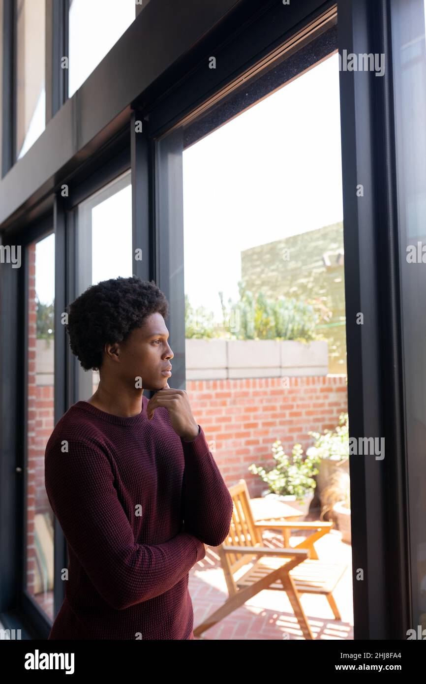 Thoughtful african american young man with hand on chin looking through window at home. unaltered, lifestyle, contemplation, day dreaming and domestic Stock Photo