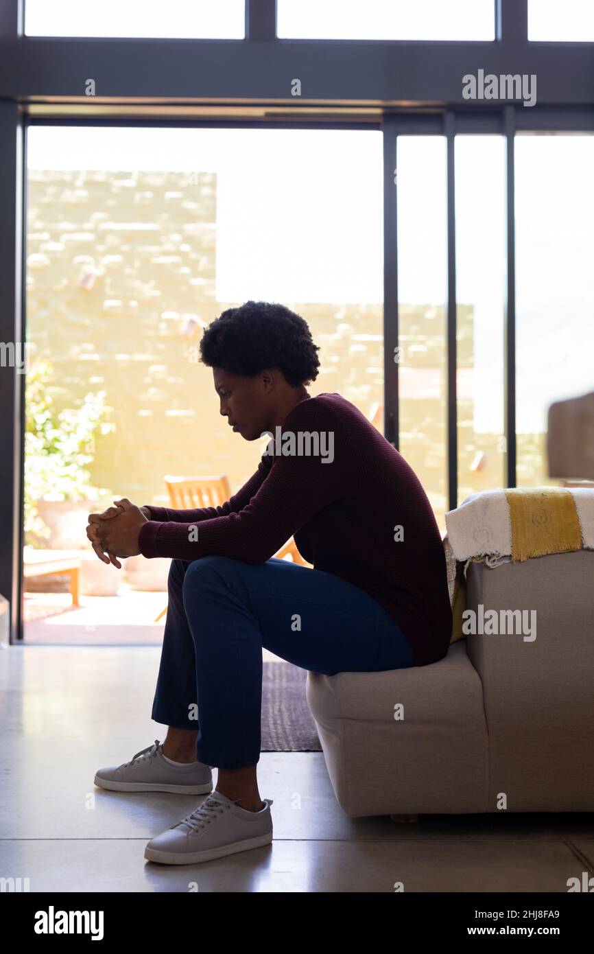 Side view of thoughtful african american young man with hands clasped sitting on sofa at home. unaltered, lifestyle, contemplation, day dreaming, depr Stock Photo