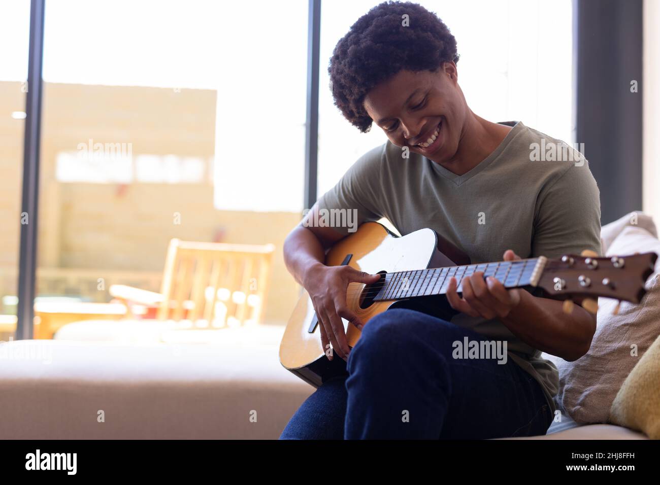 Smiling african american young man playing guitar while sitting on sofa ...