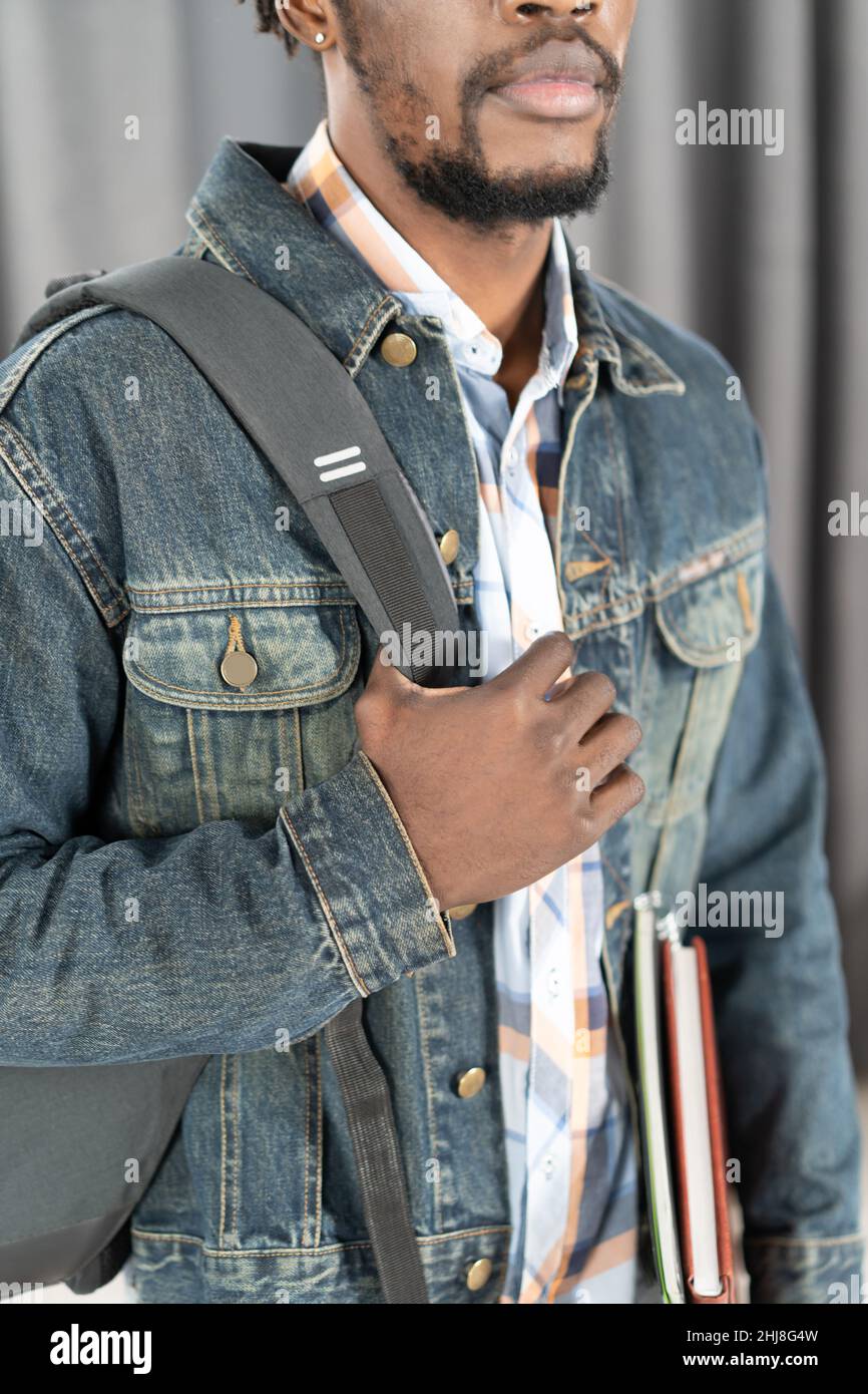 No face young african-american man with beard holding books and backpack strap wearing denim jacket isolated on grey. Handsome young student man on the way to school. School concept. Studies concept.  Stock Photo