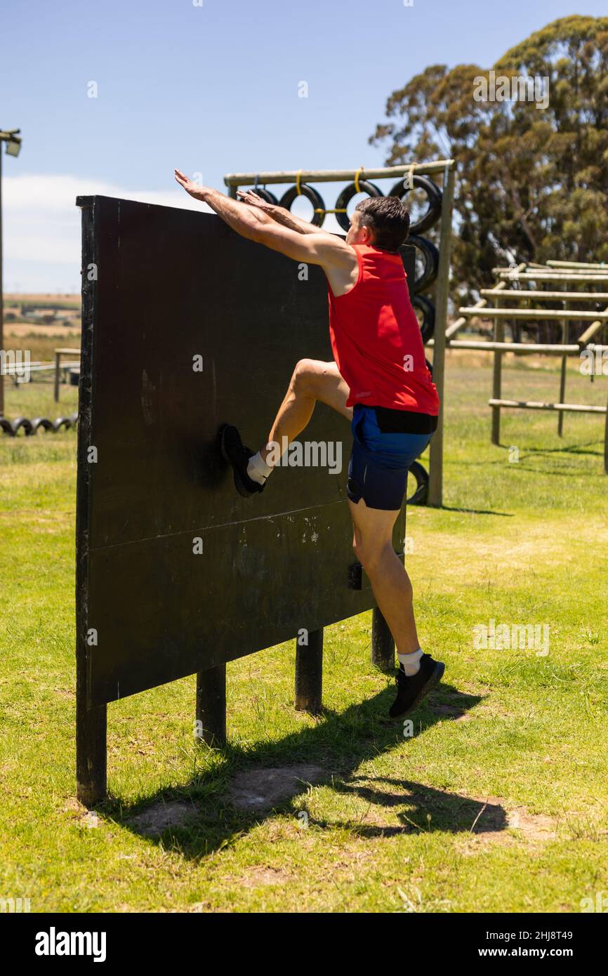 Caucasian fit man climbing a wooden wall during obstacle course at boot camp Stock Photo