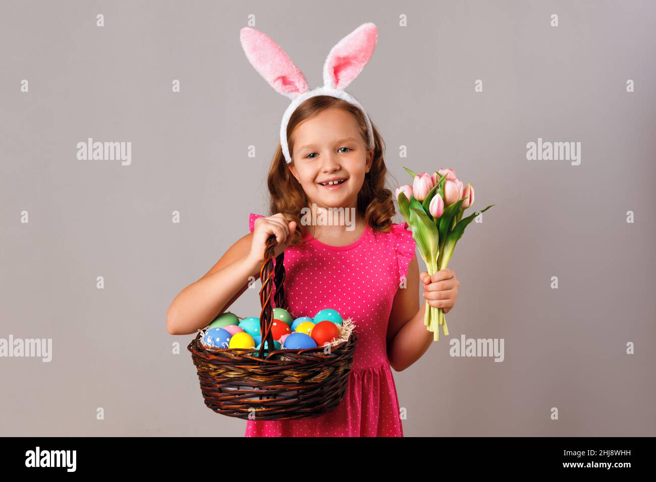 A cute little girl in easter bunny ears holds a basket of eggs and a bouquet of tulips. Portrait of a child on a gray isolated background. Stock Photo