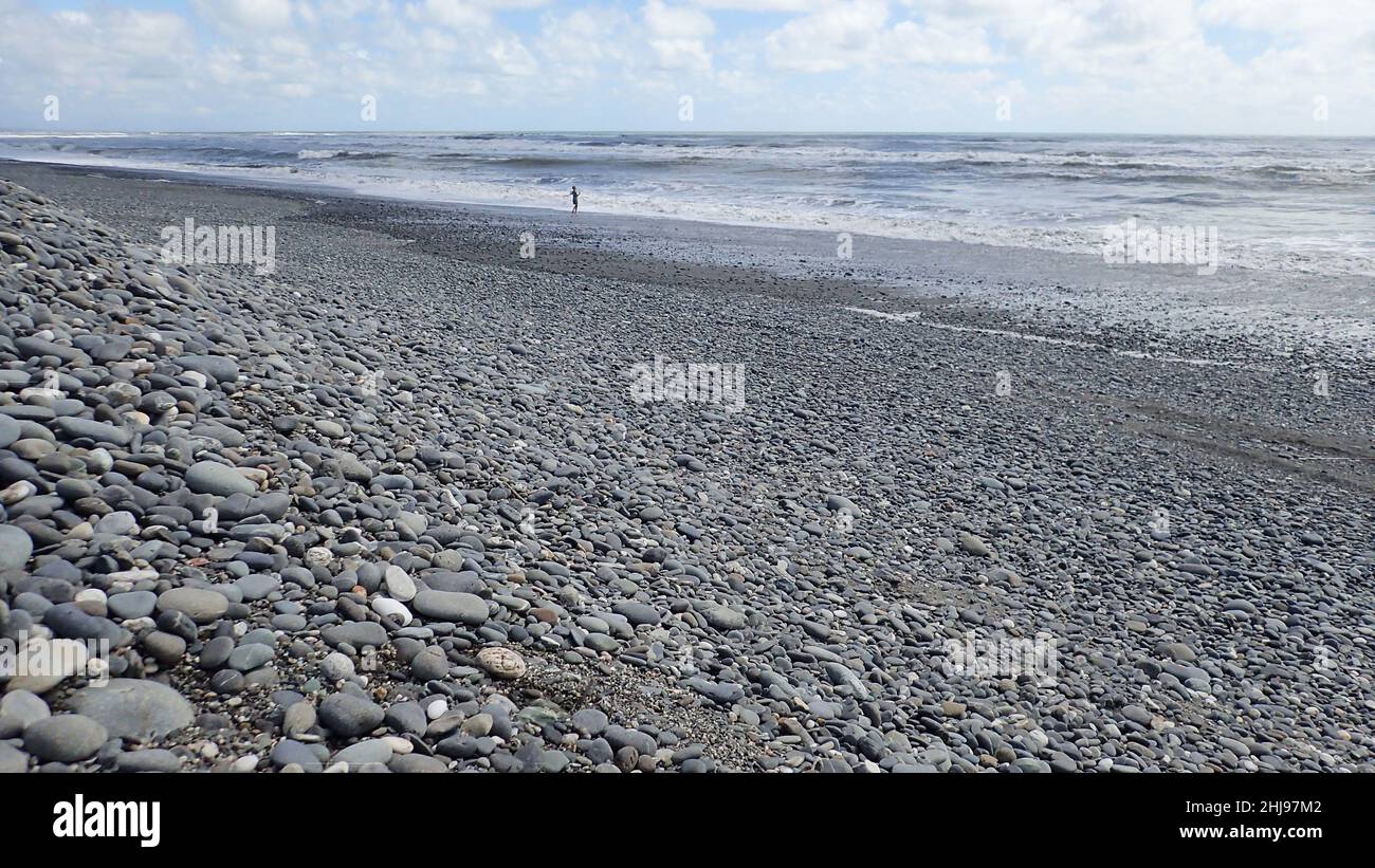 One distant person at the surf's edge on a big stony beach on NZ's West Coast Stock Photo