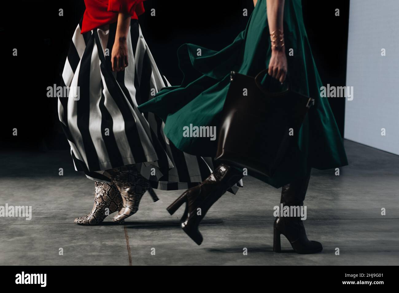 Two cropped female figures dressed in a stylish long striped black white skirt, red jacket and green dress walking the runway. Fashion week details Stock Photo