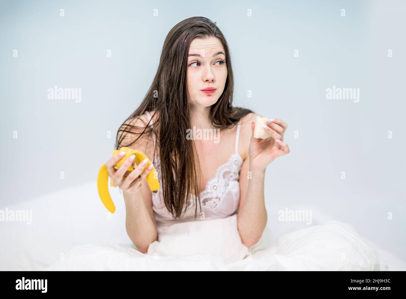 The brunette is having breakfast with fruit. The girl is sitting on the bed, wrapped in a blanket, and enjoying eating a banana. Stock Photo