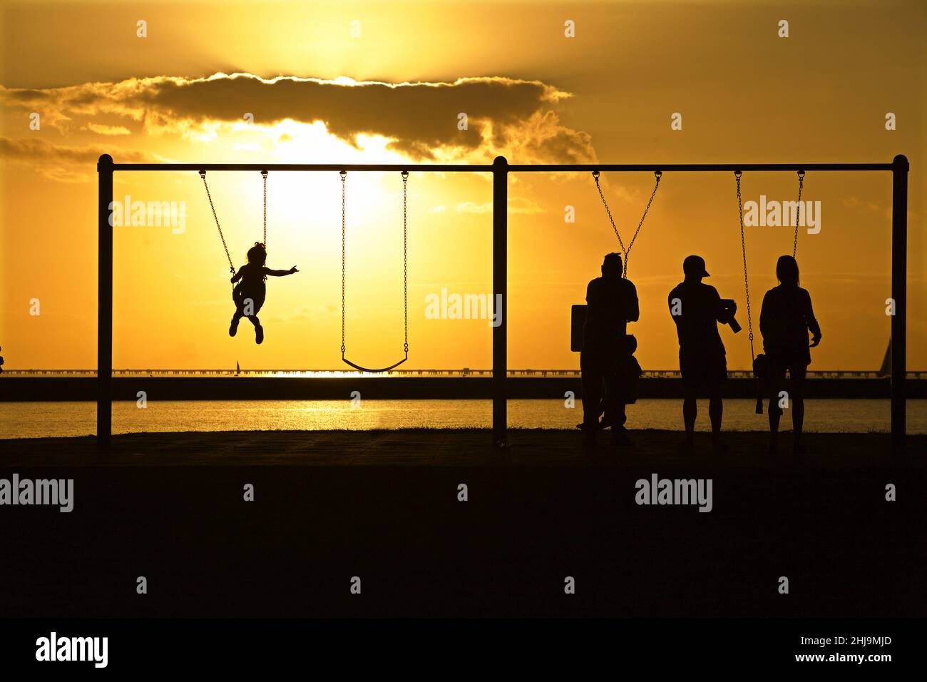 Parents and children in silhouette play on swings at sunset Stock Photo
