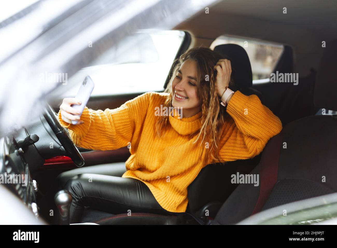 Portrait of a young casual woman in orange sweater, texting on her smartphone or making video call ,while driving or traffic stop a car during lunch break. Stock Photo