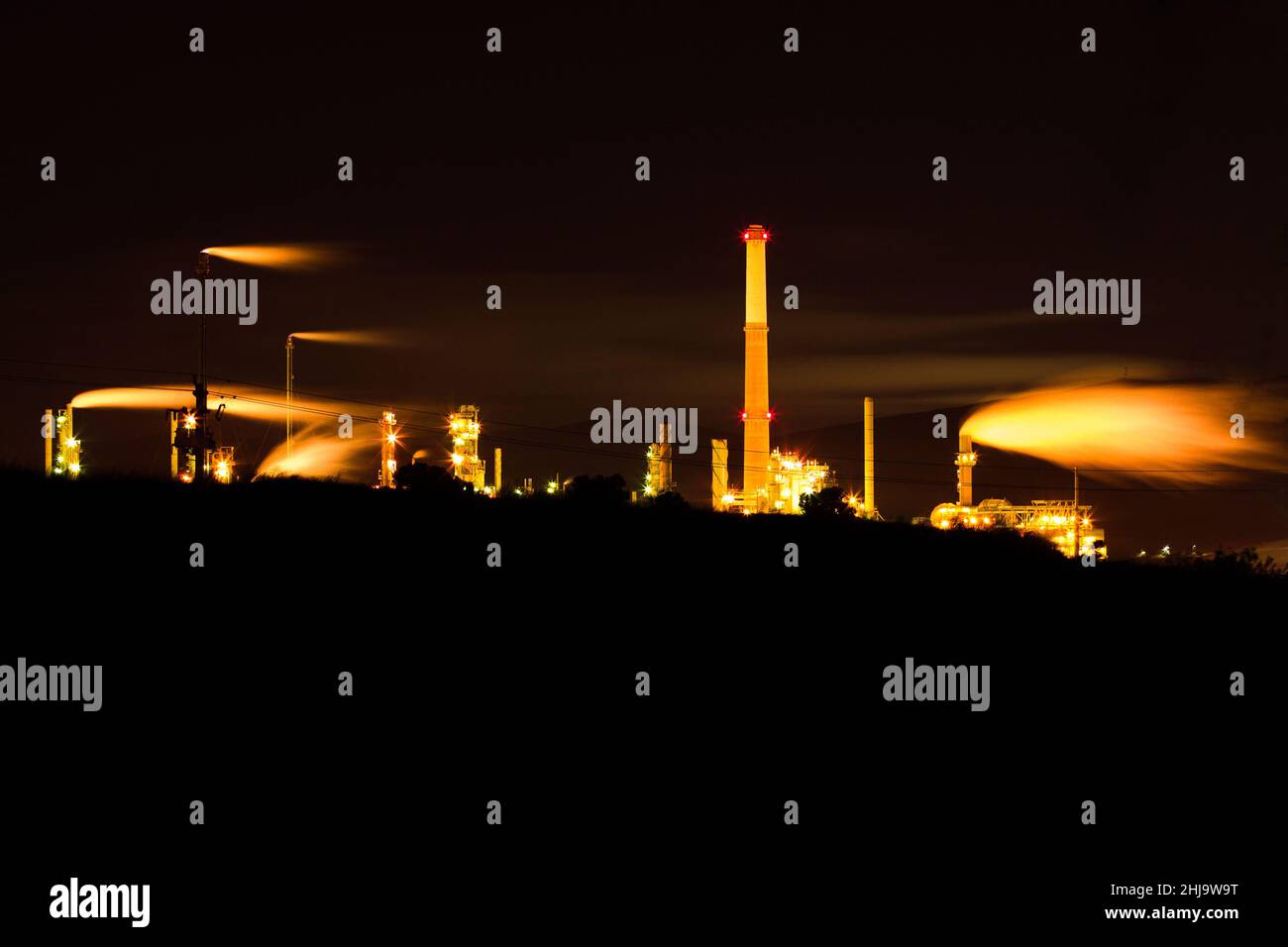 An oil refinery with glowing yellow smoke pollution rising into the dark black night sky. Stock Photo