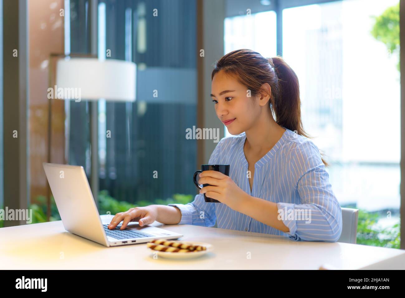Asian woman drinking coffee in mug and smiling happily while taking a break from work at her computer during work at home in the living room. Stock Photo