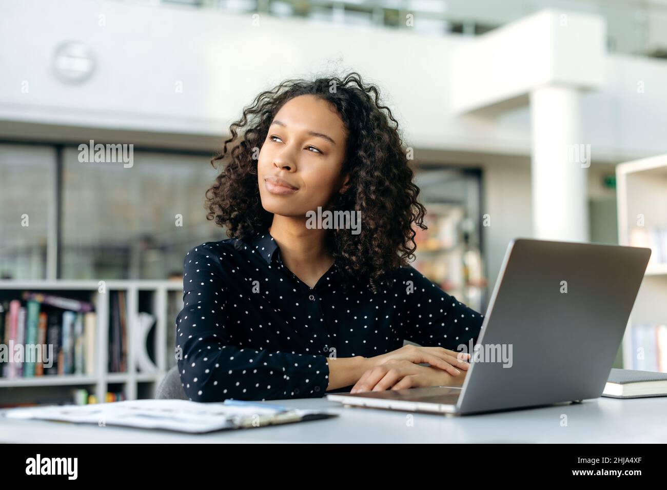 Positive lovely african american girl, office worker, sales manager, corporate marketer, sit at work desk in modern office, looking away, planning strategy, thinking project, dreaming about vacation Stock Photo