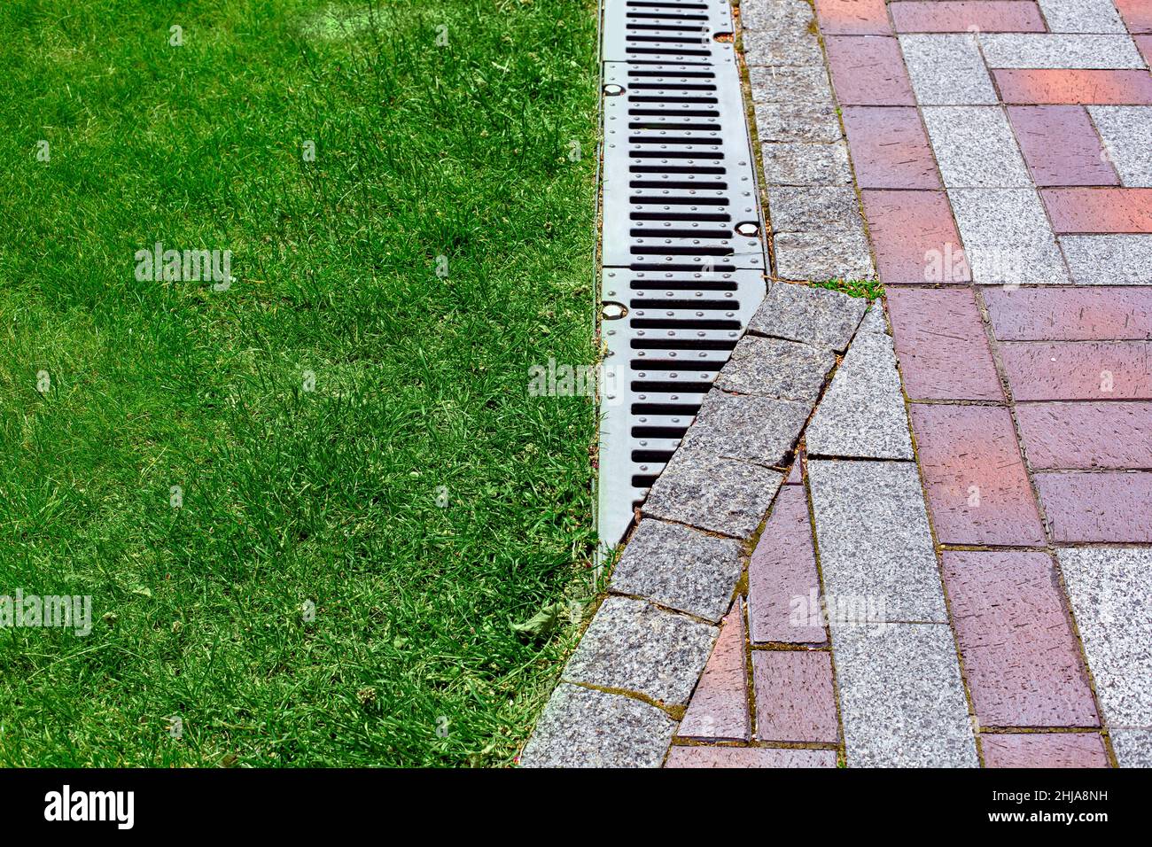 drainage grate bolted to storm drain at corner of pedestrian pavement walk way made of stone brick tiles gray and red pattern in backyard with copy sp Stock Photo