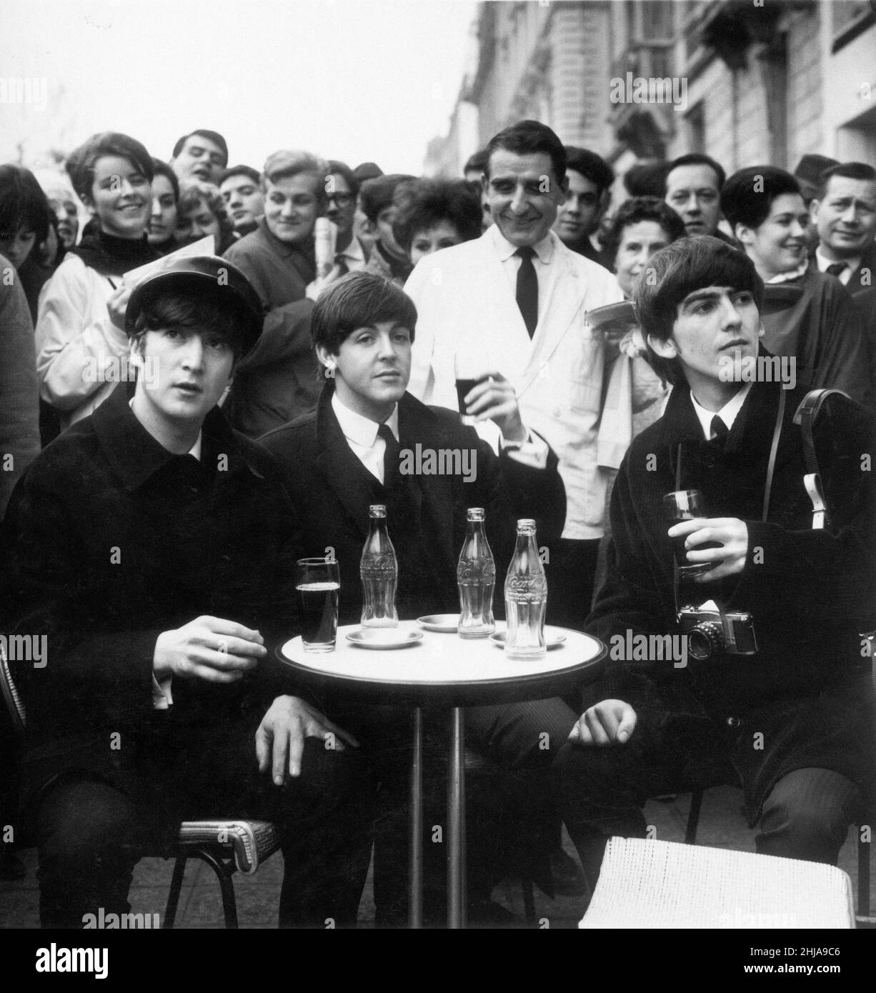 Three Beatles - John, Paul and George enjoy a glass of coca cola at a street cafe in Paris France 15th January 1964.  George is carrying his Pentax Camera.  The Beatles performed at the Olympia Theatre Paris, continuing their 18 date residency which began on 16 January 1964 and ended on 4 February.   It was in Paris, that they learnt that they were number one in the American charts, which prompted a visit to New York the following week, where they conquered America first appearing on The Ed Sullivan Show.  Picture taken 15th January 1964 Stock Photo