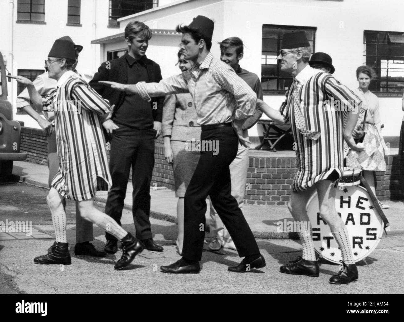 Recognise the hatted 'busker'? It's Cliff Richard, doing a soft-shoe shuffle with London's Road Stars buskers. Cliff had invited them to Elstree Studios where he is filming Summer Holiday to register his own protest at the recent ban on the buskers. Later, the buskers went off to rehearse for an engagement in Westminster. Asked Cliff: 'Aren't you scared of getting pinched for obstruction?' Said the Old 'Uns to the Young One: 'Not a chance. We're performing at a cop's wedding.' August 1962 P014470 Stock Photo