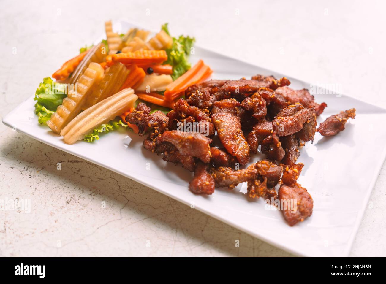 Close up deep-fried pork with sesame in beautiful white plate, side dish with pickled vegetables and fresh vegetable on a white stone table, Thai food Stock Photo