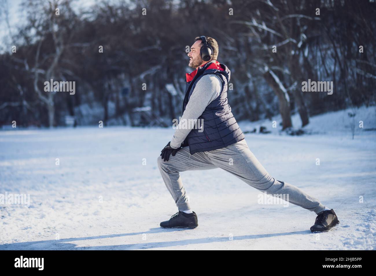 Adult man is exercising in park in wintertime. He is stretching his body before jogging. Stock Photo