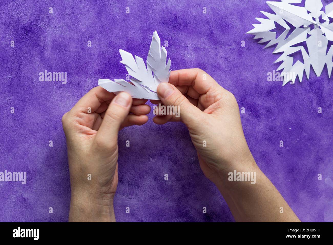 Woman hands unfolding paper to reveal snowflake on violet surface Stock Photo