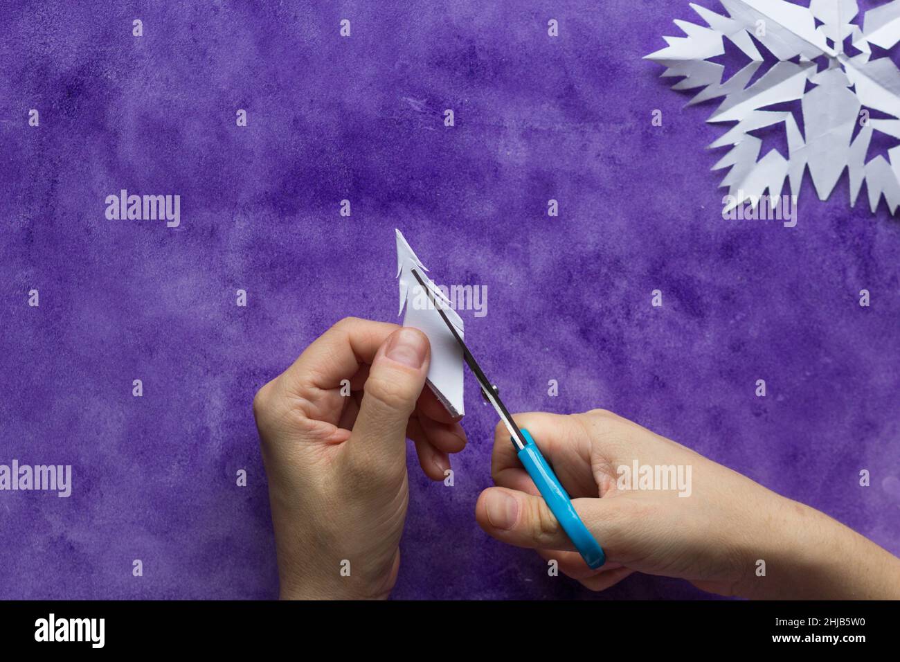 Woman hands cutting away shapes from the edges of the paper to shape snowflake pattern on violet surface Stock Photo