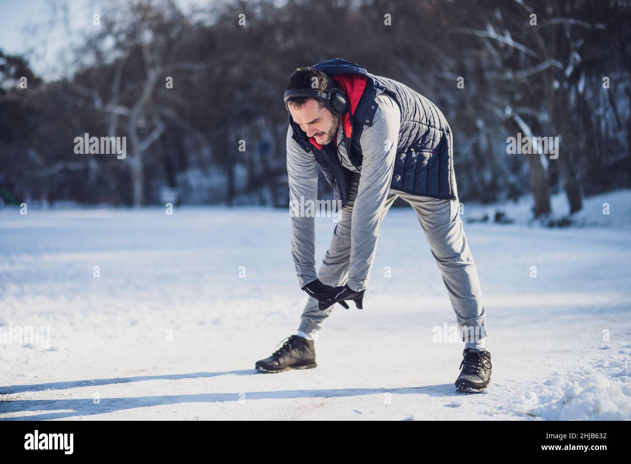 Adult man is exercising in park in wintertime. He is stretching his body before jogging. Stock Photo