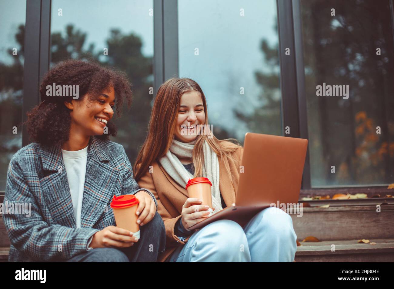 Two happy positive multi-ethnic teen girls with computer and drinking take away coffee outdoors, black and white female best friends looking at laptop Stock Photo