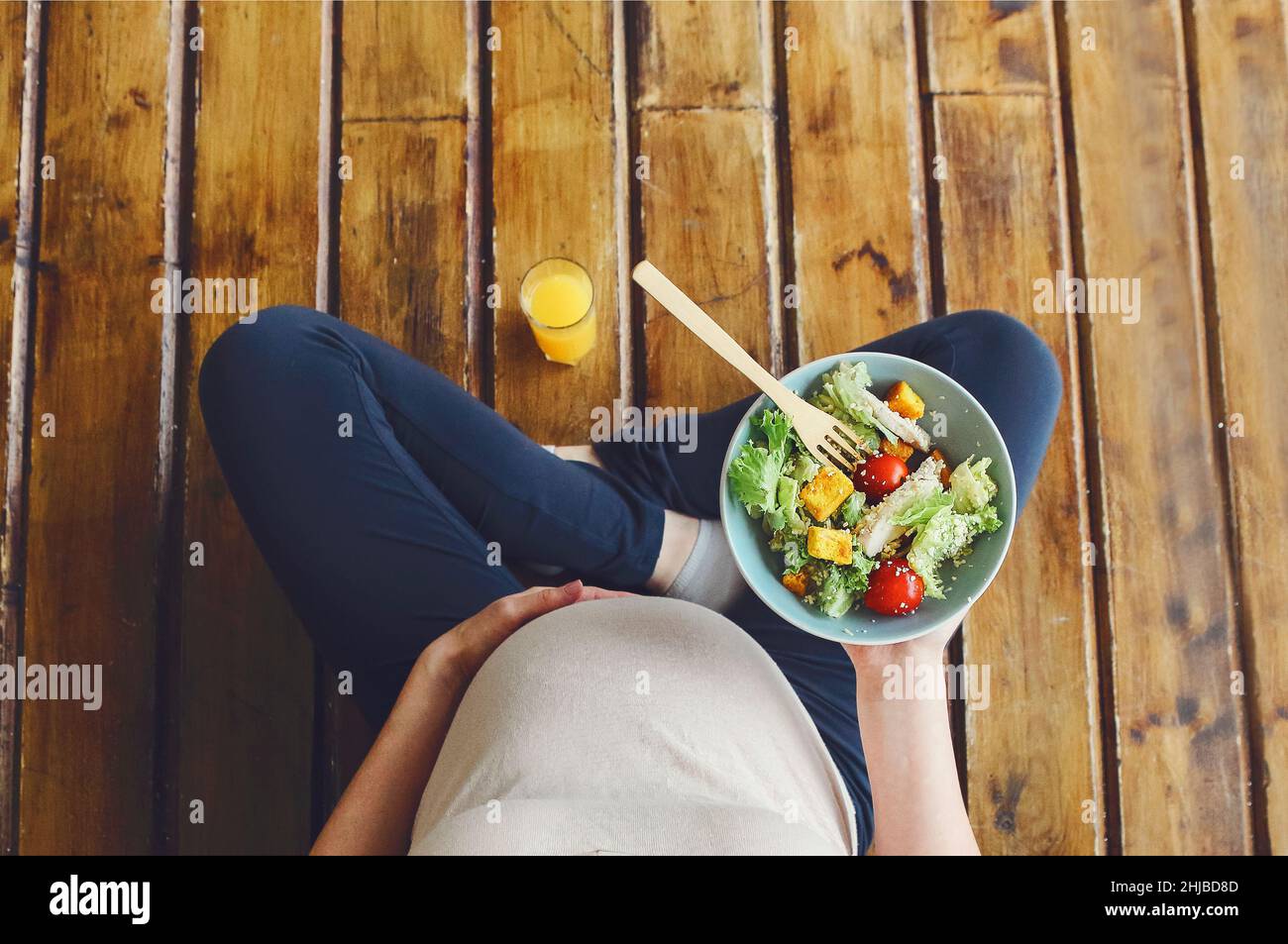 View from above of pregnant woman with fresh salad bowl and orange juice, expectant mother having healthy lunch while sitting on wooden floor in lotus Stock Photo