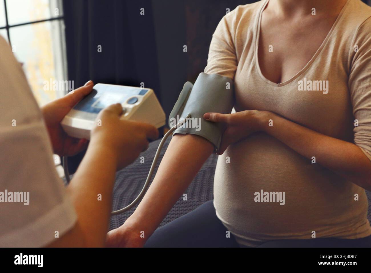 Doctor measuring blood pressure of pregnant woman while visiting her at home, therapist examining expectant mother during prenatal care appointment in Stock Photo