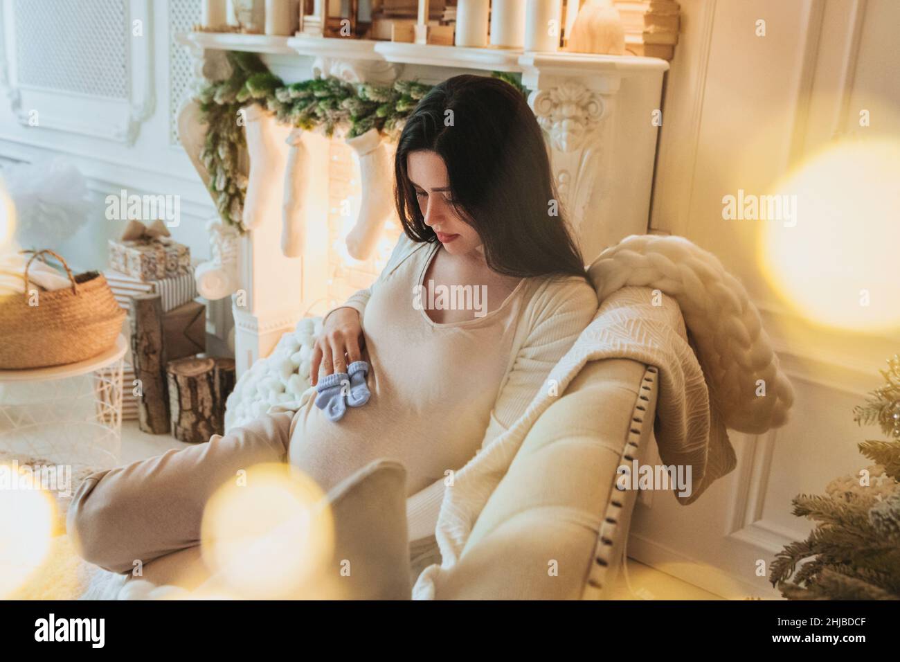 Young pregnant woman with pair of cute blue baby socks relaxing near fireplace at home during Christmas time. Relaxed expectant mother resting on sofa Stock Photo