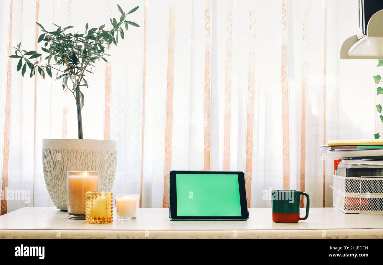 Digital tablet with green screen on wooden table with houseplant, candles, cup of tea and office supplies, cozy remote workplace with touch device. Mo Stock Photo